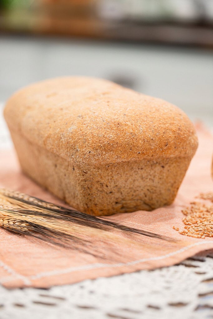 Fresh milled multigrain seeded sourdough sandwich bread loaf with a hearty textured crust resting on a peach linen, beside dried wheat stalks and scattered sesame seeds, with a soft-focus neutral background.