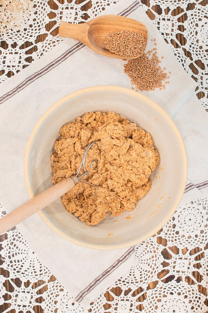 Overhead view of fresh milled multigrain seeded sourdough bread dough being mixed in a cream ceramic bowl with a Danish dough whisk, with whole wheat berries spilling from a wooden scoop on a white striped linen with crochet lace.