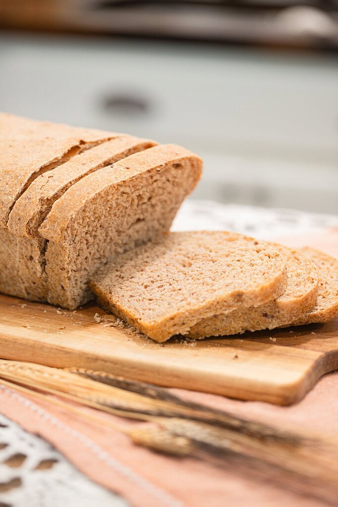 Close-up of a sliced fresh milled multigrain seeded sourdough sandwich bread loaf on a wood cutting board, showing a dense, hearty crumb with three slices fanned forward, with dried wheat stalks alongside on a peach linen and soft-focus kitchen background.