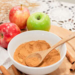 A white bowl of blended homemade apple pie spice with a wooden spoon, surrounded by red and green apples, cinnamon sticks, and whole nutmeg on a wood cutting board with a lace doily.