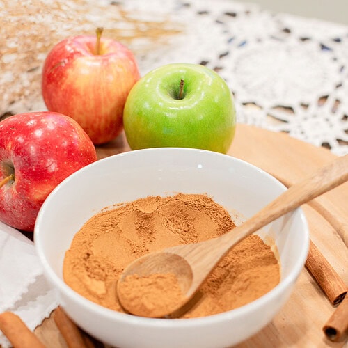A white bowl of blended homemade apple pie spice with a wooden spoon, surrounded by red and green apples, cinnamon sticks, and whole nutmeg on a wood cutting board with a lace doily.