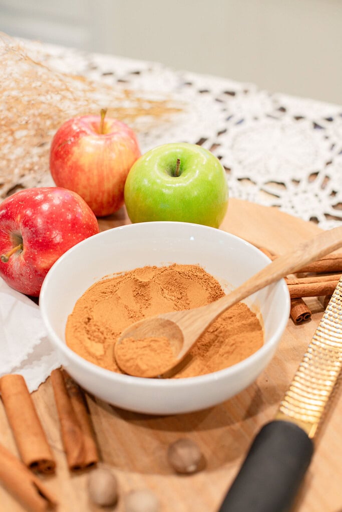 A white bowl of blended apple pie spice with a wooden spoon on a wood cutting board, styled with red and green apples, cinnamon sticks, whole nutmeg, and a fine grater alongside a lace doily.