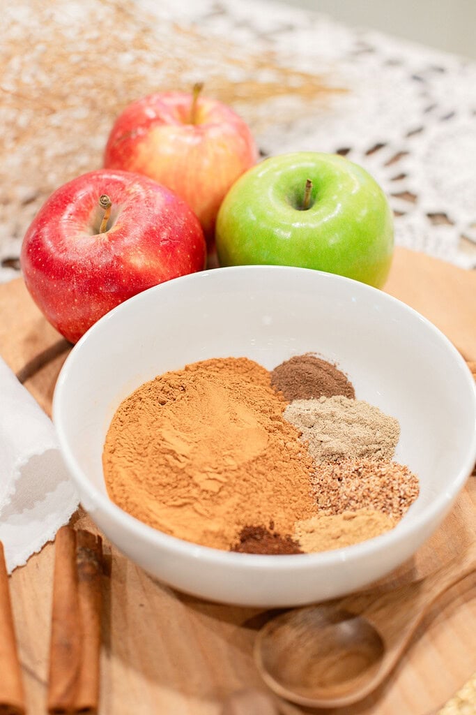 A white bowl showing the individual measured spices for homemade apple pie spice blend before mixing, with cinnamon, nutmeg, cardamom, and allspice portioned separately, styled with red and green apples and cinnamon sticks on a wood cutting board.
