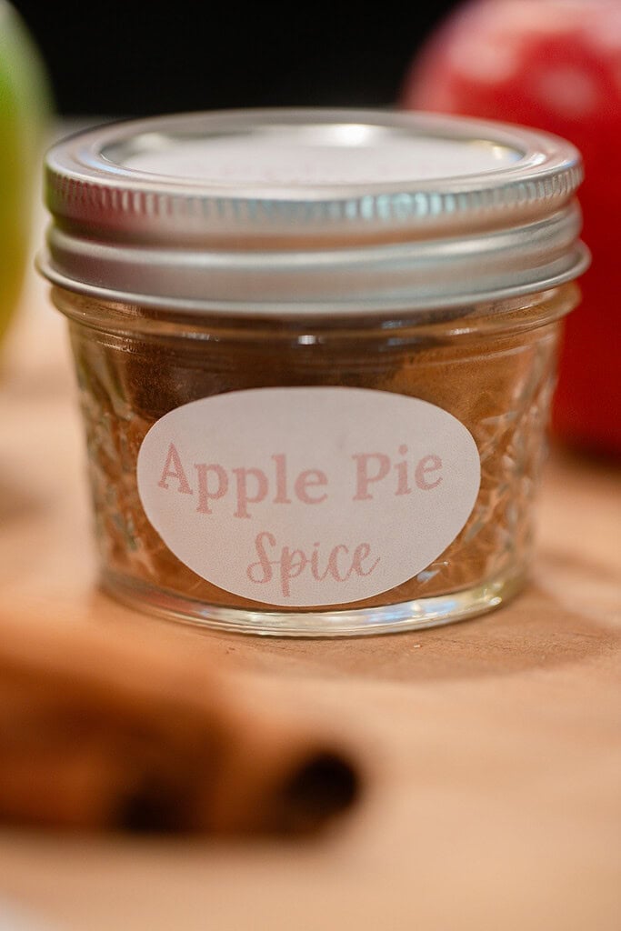 A small glass jar filled with homemade apple pie spice blend and sealed with a silver lid, labeled with a handwritten Apple Pie Spice tag, with cinnamon sticks and apples softly blurred in the background.