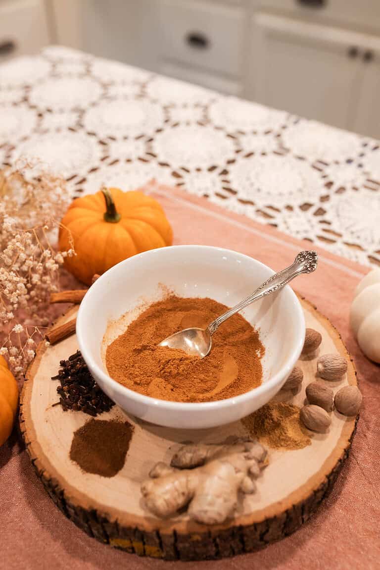 A whie ceramic bowl with a spice blend for Pumpkin Pie Seasoning with the whole seasoning around the bowl on a slice of wood with a pumpkin beside the bowl on a wooden counter top with a crochet tablecloth in a cottage kitchen.