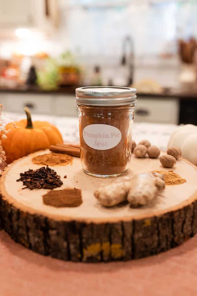 A jar of Pumpkin Pie Spice on a slice of a log on a kitchen tea towel with a pumpkin beside it and the seasonings all around the jar in a cottage kitchen.