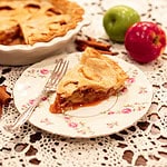 A detailed view of a slice of apple pie on a floral plate, showing the rich apple filling and buttery, flaky crust, with green and red apples and cinnamon sticks in the background.