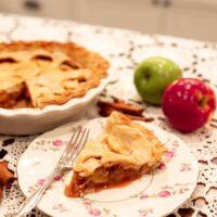 A detailed view of a slice of apple pie on a floral plate, showing the rich apple filling and buttery, flaky crust, with green and red apples and cinnamon sticks in the background.