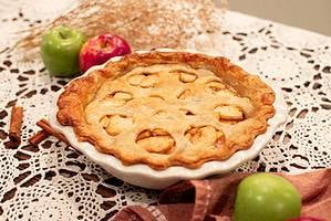 A whole homemade apple pie in a ceramic pie dish with a golden, lattice-like crust. The pie is surrounded by red and green apples, cinnamon sticks, and dried floral decor, all set on a lace tablecloth.