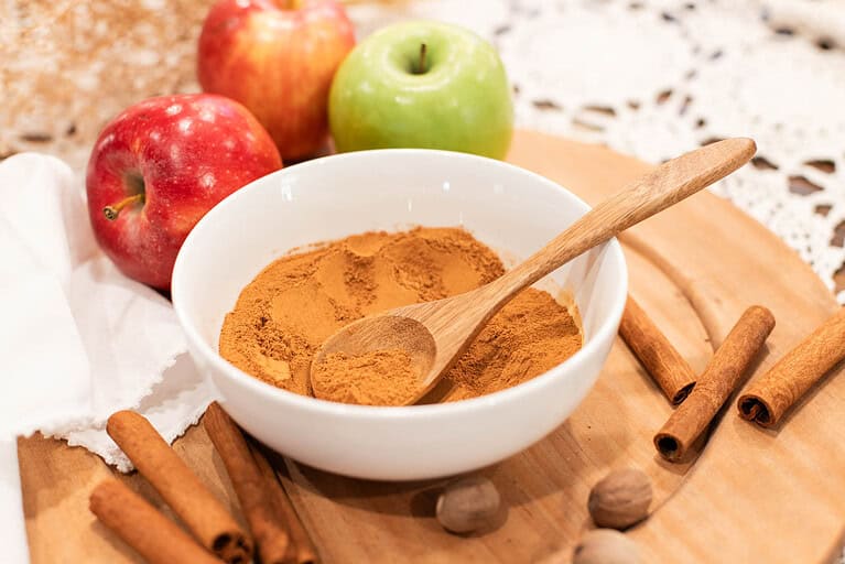 A bowl of homemade apple pie spice surrounded by fresh red and green apples, cinnamon sticks, and nutmeg on a wooden board, perfect for fall baking.