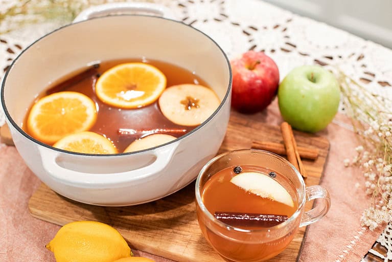 A pot of mulled cider and a mug full of it with fresh apples, oranges amd cinnamon sticks scattered around both in a cottage kitchen on a crochet tablecloth.