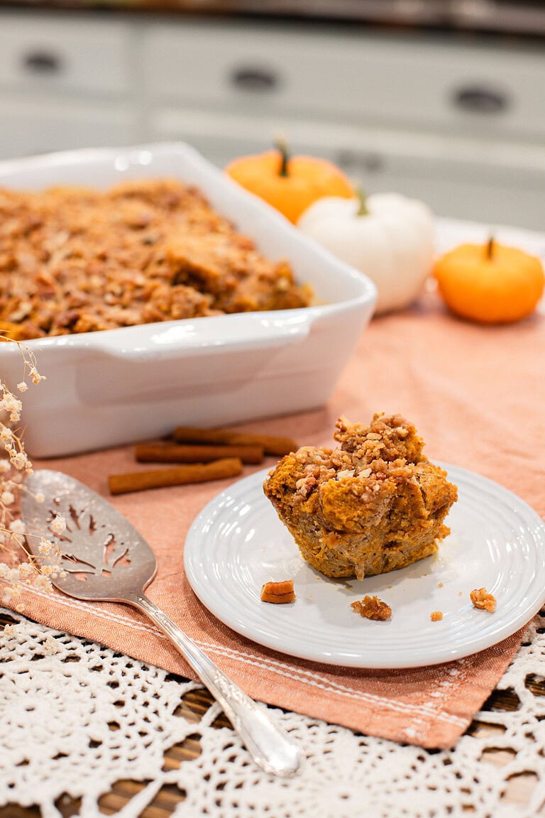 Single serving of overnight sourdough pumpkin French toast casserole with brown sugar pecan streusel on a white plate beside a decorative serving spatula, with the full casserole dish and mini pumpkins in the background