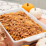 White baking dish of sourdough pumpkin French toast casserole topped with golden brown sugar pecan streusel, surrounded by orange and white mini pumpkins and cinnamon sticks on a peach linen