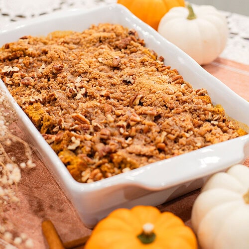 White baking dish of sourdough pumpkin French toast casserole topped with golden brown sugar pecan streusel, surrounded by orange and white mini pumpkins and cinnamon sticks on a peach linen