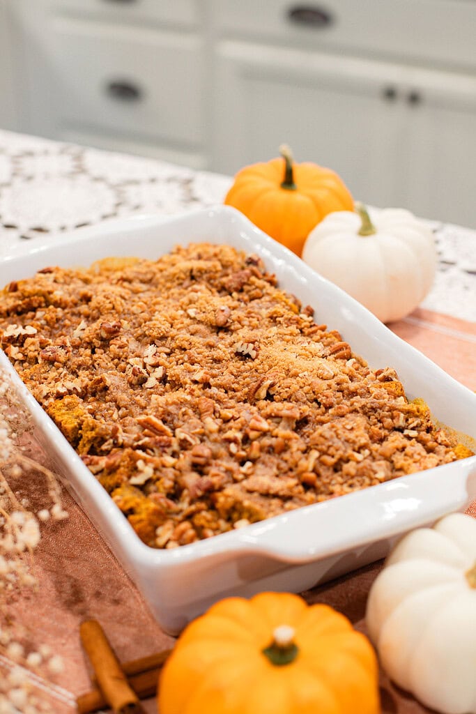 White baking dish of sourdough pumpkin French toast casserole topped with golden brown sugar pecan streusel, surrounded by orange and white mini pumpkins and cinnamon sticks on a peach linen