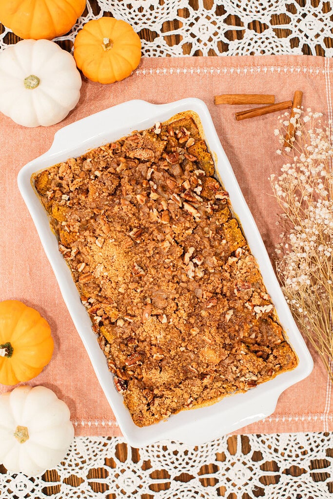Overhead flat lay of a full baked sourdough pumpkin French toast casserole with pecan brown sugar streusel in a white baking dish, surrounded by orange and white mini pumpkins, cinnamon sticks, and dried baby's breath on a peach linen