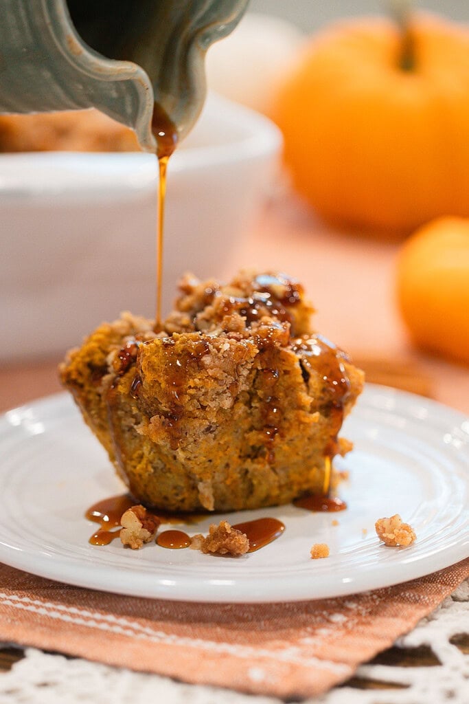 Maple syrup being poured from a ceramic pitcher over a single serving of sourdough pumpkin French toast casserole with pecan streusel on a white plate, with mini pumpkins in the soft background