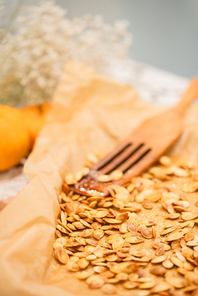 Close-up of golden roasted pumpkin seeds spread on parchment paper with a wooden slotted spoon, styled with a mini pumpkin and dried baby's breath in the background.