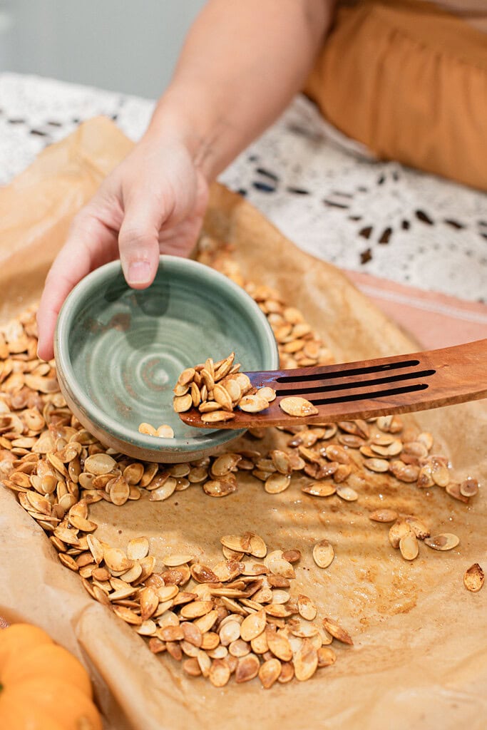 A hand scooping freshly roasted pumpkin seeds from a parchment-lined baking sheet into a small sage green ceramic bowl using a wooden slotted spoon.