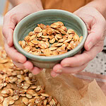 Two hands cupping a sage green ceramic bowl filled with golden roasted pumpkin seeds, with more seeds visible on a parchment-lined baking sheet below.