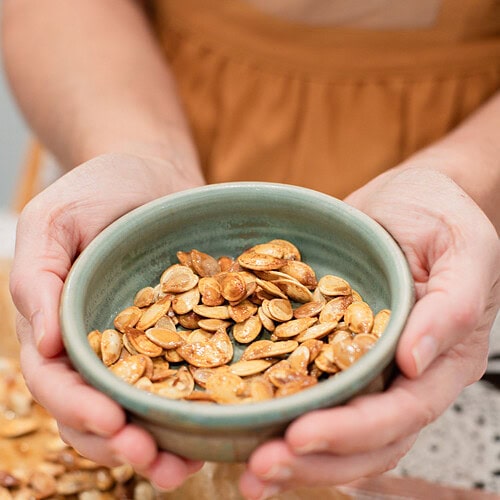 Two hands cupping a sage green ceramic bowl filled with golden roasted pumpkin seeds, with more seeds visible on a parchment-lined baking sheet below.