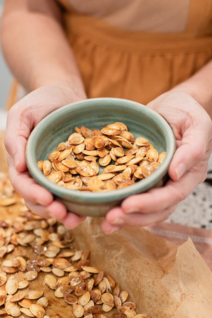 Two hands cupping a sage green ceramic bowl filled with golden roasted pumpkin seeds, with more seeds visible on a parchment-lined baking sheet below.
