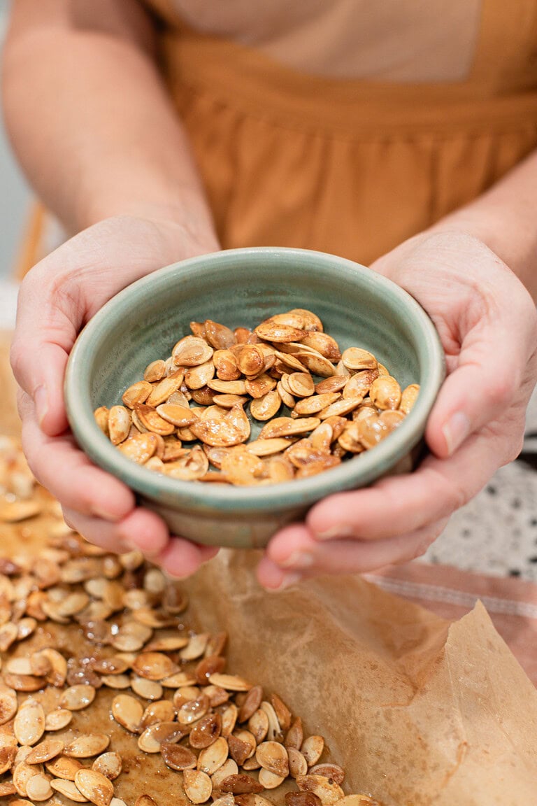 Two hands cupping a sage green ceramic bowl filled with golden roasted pumpkin seeds, with more seeds visible on a parchment-lined baking sheet below.