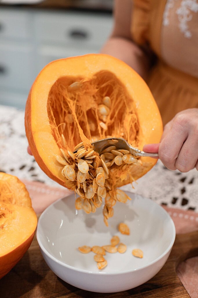 A hand scooping raw pumpkin seeds from the hollow of a freshly cut pumpkin half into a white bowl, showing the stringy orange interior and seeds falling into the bowl below.