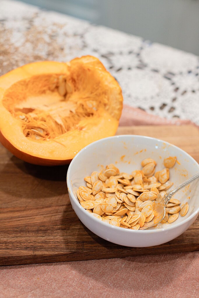 A white bowl filled with raw pumpkin seeds next to a hollowed-out pumpkin half on a wooden cutting board, styled on a dusty rose linen with a lace doily.