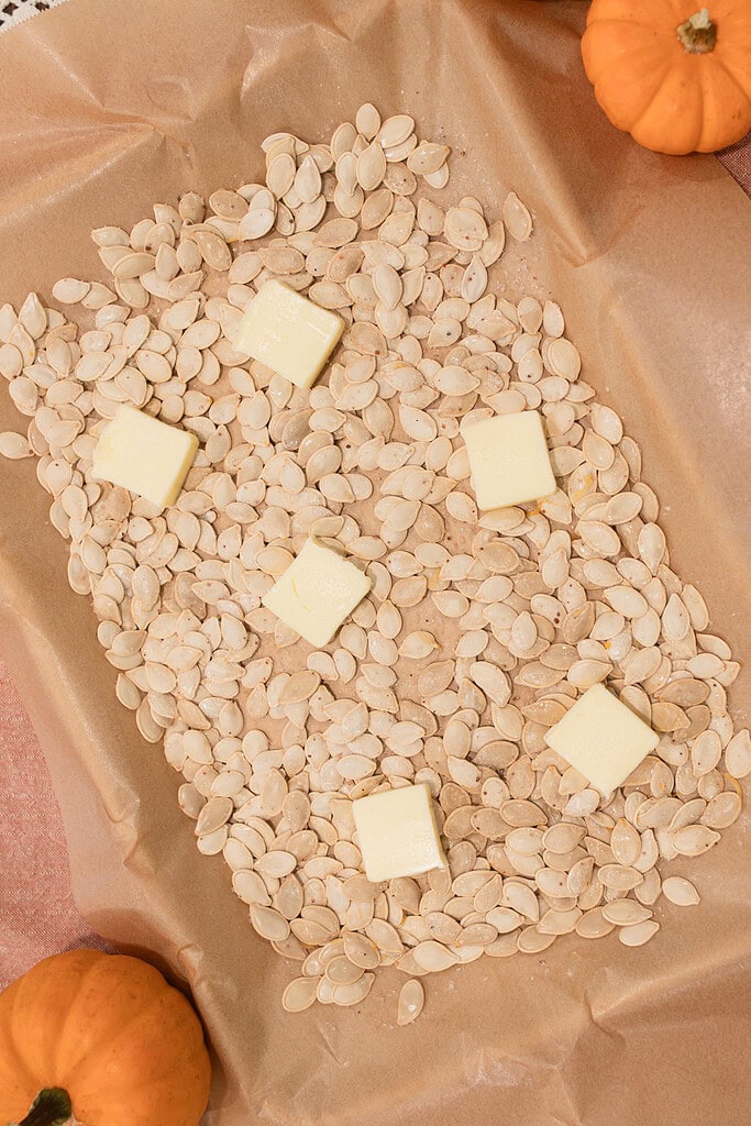 Overhead view of raw pumpkin seeds spread on a parchment-lined baking sheet with pats of butter placed on top before roasting, with mini orange pumpkins in the corners.