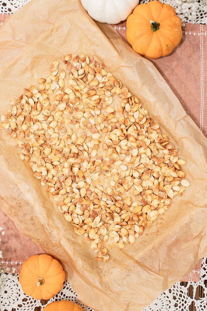 Overhead view of roasted pumpkin seeds spread in a single layer on a parchment-lined baking sheet, styled with mini orange and white pumpkins on a dusty rose linen.