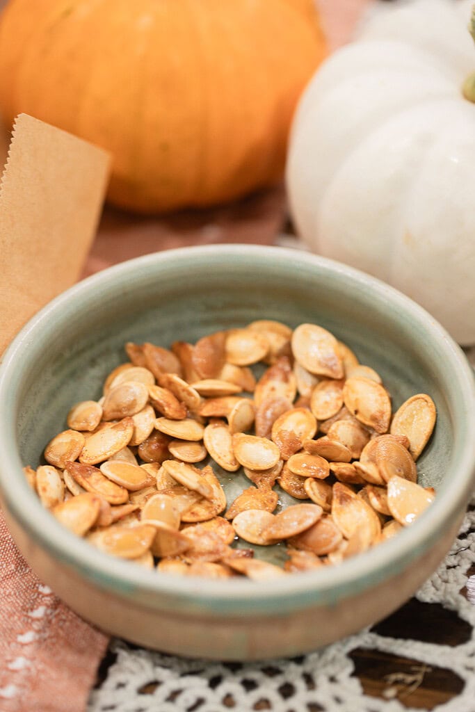 A sage green ceramic bowl filled with golden roasted pumpkin seeds, styled with an orange and a white pumpkin softly blurred in the background on a dusty rose linen.