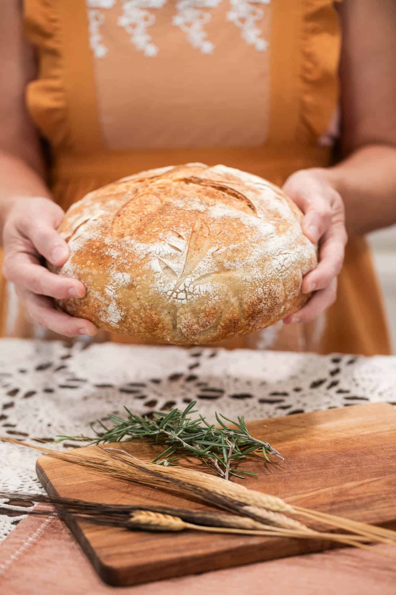 Hands holding a warm loaf of rosemary sourdough bread, showing off its rustic and artisanal design with a floral scoring pattern.