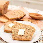 A cozy scene of two slices of buttered multigrain sourdough bread on a white plate, with a sliced loaf of bread and wheat stalks in the background.