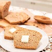 A cozy scene of two slices of buttered multigrain sourdough bread on a white plate, with a sliced loaf of bread and wheat stalks in the background.