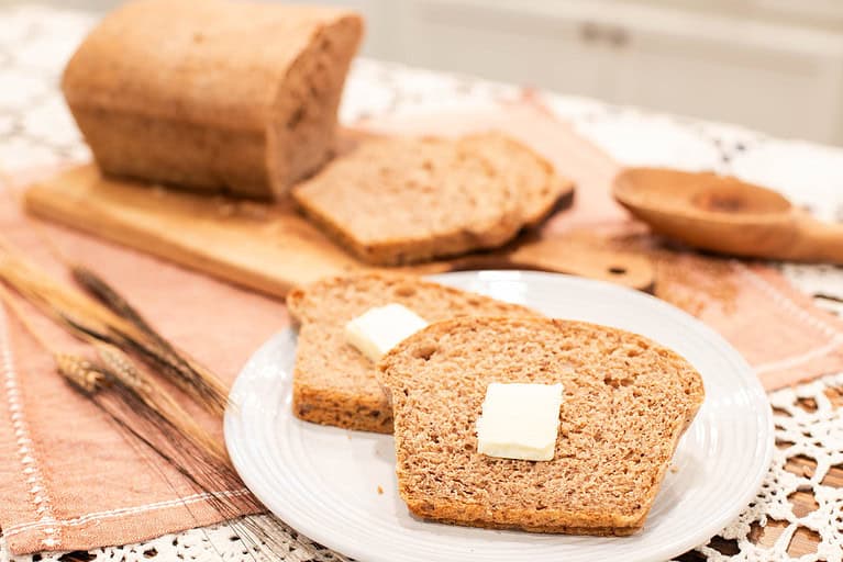 A cozy scene of two slices of buttered multigrain sourdough bread on a white plate, with a sliced loaf of bread and wheat stalks in the background.