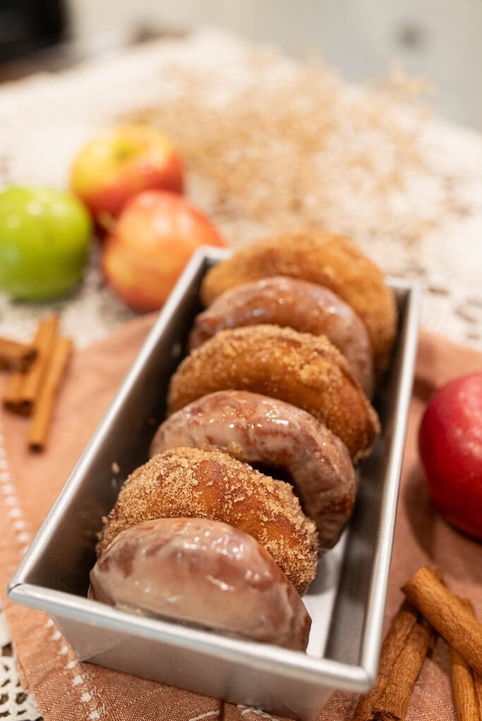 Sourdough apple cider donuts arranged in a loaf pan, coated in cinnamon sugar and glaze with apples in the background