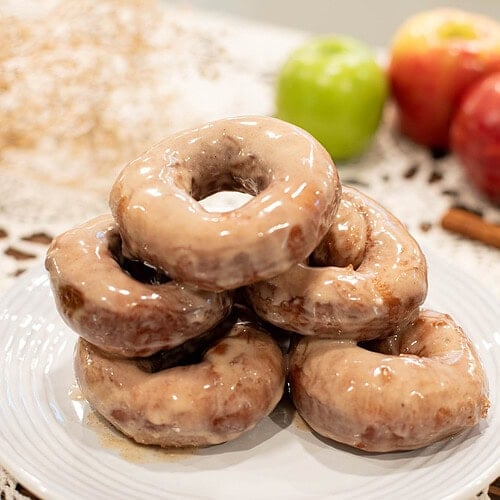 Stack of glazed sourdough apple cider donuts on a white plate with apples in the background