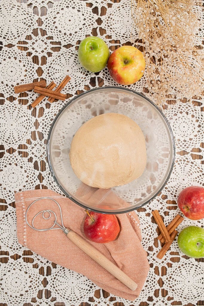 Smooth sourdough donut dough resting in a glass bowl with apples and cinnamon sticks on a lace tablecloth