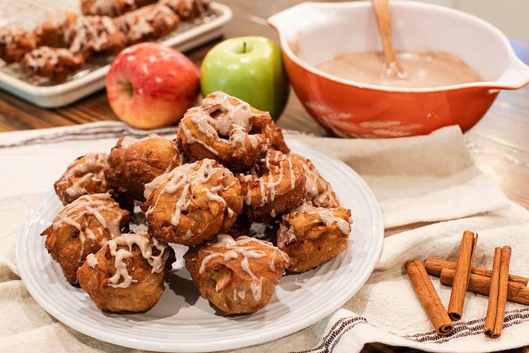 A plate full of freshly made sourdough apple fritters with more in the background and a bowl of glaze in a cottage kitchen.