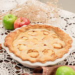 A whole baked sourdough apple pie with apple-shaped cutouts in the top crust in a white ceramic pie dish, styled on a lace doily with red and green apples, dried botanicals, and a dusty rose linen.