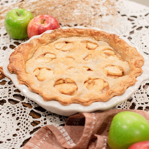 A whole baked sourdough apple pie with apple-shaped cutouts in the top crust in a white ceramic pie dish, styled on a lace doily with red and green apples, dried botanicals, and a dusty rose linen.