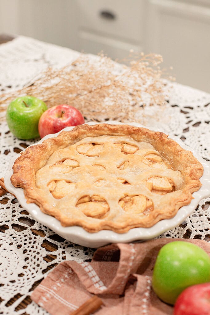 A whole baked sourdough apple pie with apple-shaped cutouts in the top crust in a white ceramic pie dish, styled on a lace doily with red and green apples, dried botanicals, and a dusty rose linen.