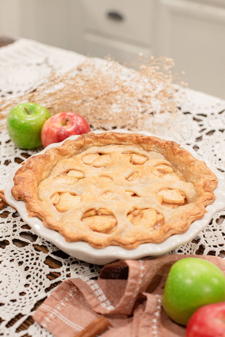 A whole baked sourdough apple pie with apple-shaped cutouts in the top crust in a white ceramic pie dish, styled on a lace doily with red and green apples, dried botanicals, and a dusty rose linen.