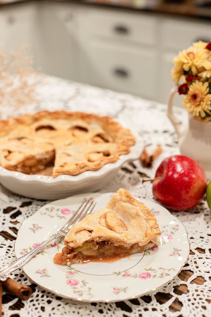 A slice of sourdough apple pie with a golden flaky crust and cinnamon apple filling on a vintage floral china plate with a silver fork, with the whole pie, a red apple, and yellow flowers softly blurred behind it.