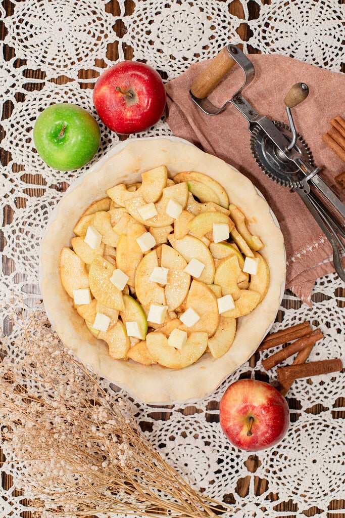 Overhead view of a raw sourdough pie shell filled with cinnamon-spiced apple slices and dotted with butter pats, styled on a lace doily with red and green apples, cinnamon sticks, dried botanicals, and a vintage pastry cutter.