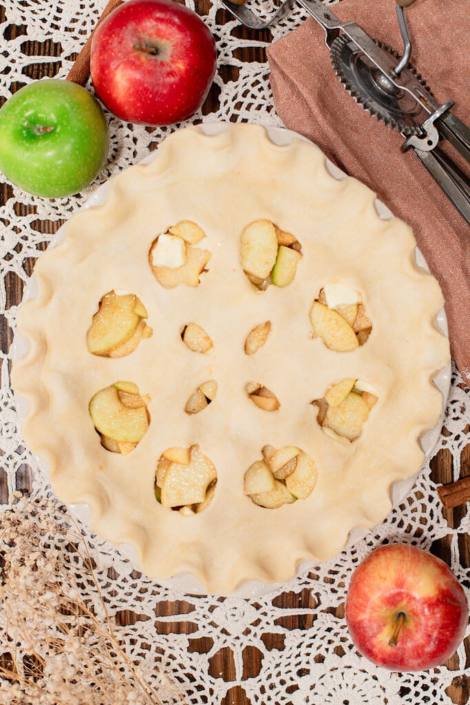 Overhead view of an unbaked sourdough apple pie with apple-shaped cutouts in the top crust revealing the spiced apple filling beneath, in a white ceramic pie dish on a lace doily with red and green apples and a vintage pastry cutter.