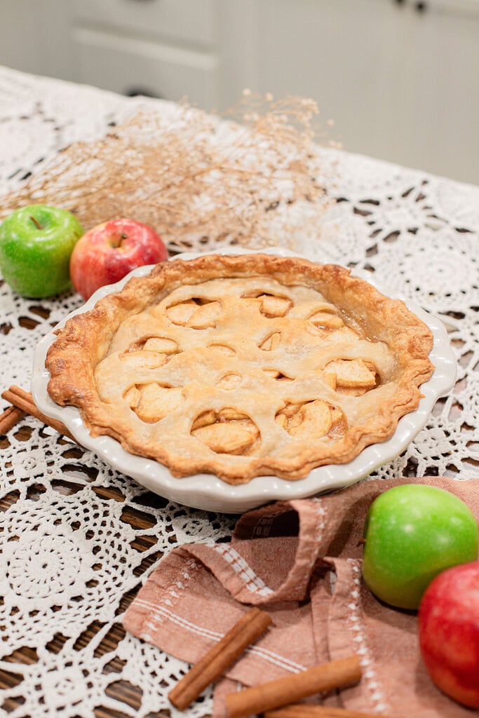 A fully baked sourdough apple pie with golden apple cutout crust in a white ceramic pie dish, styled on a lace doily with green and red apples, cinnamon sticks, dried botanicals, and a dusty rose linen in a cottage kitchen.