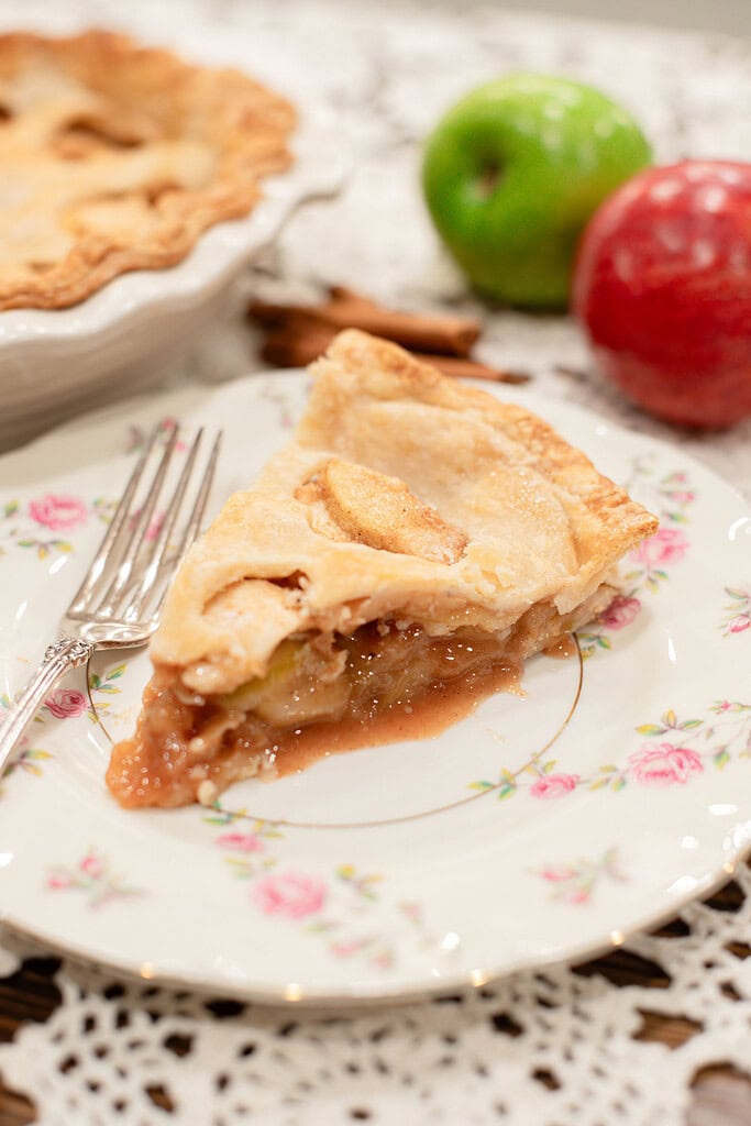 A close-up slice of sourdough apple pie showing the flaky layered crust and juicy cinnamon apple filling on a vintage floral china plate with a silver fork, with a green apple, red apple, and the whole pie softly blurred behind it.