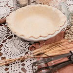 An unbaked sourdough pie crust fitted into a white ceramic pie dish with a crimped ruffled edge, surrounded by a vintage flour sifter, a glass jar of dried beans for blind baking, dried grain stalks, and vintage pastry tools on a crocheted doily over dark wood.